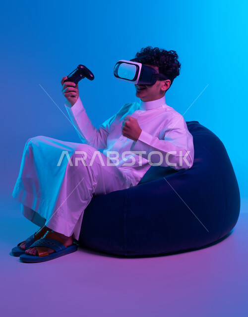 التسلية في أوقات الفراغ  بالفيديو جيمز، العالم الرقمي الترفيهي، تجربة مثيرة في الألعاب الالكترونية والأجهزة الحديثة، بورتريه لشاب عربي خليجي سعودي يرتدي الثوب التقليدي ونظارة في آر يجلس على كرسي بين باج و يمسك يد التحكم ويرفع قبضته بإيماءات النصر، خلفية زرقاء