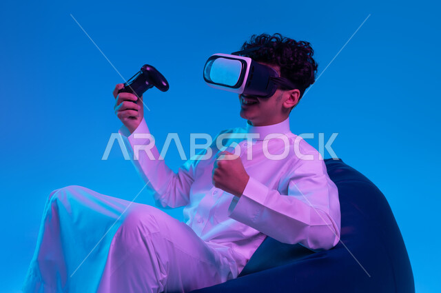 تجربة مثيرة في الألعاب الالكترونية والأجهزة الحديثة، التسلية في أوقات الفراغ  بالفيديو جيمز، العالم الرقمي الترفيهي، بورتريه لشاب عربي خليجي سعودي يرتدي الثوب التقليدي ونظارة في آر يجلس على كرسي بين باج و يمسك يد التحكم ويرفع قبضته بإيماءات النصر، خلفية زرقاء