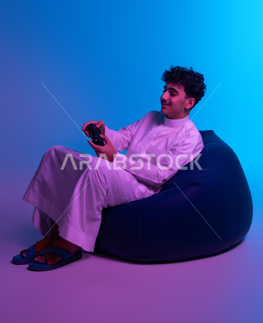 الاستمتاع باللعب بالألعاب الالكترونية الترفيهية، استخدام الأجهزة التكنولوجية والتقنيات الحديثة، بورتريه لشاب عربي خليجي سعودي مبتسم يرتدي الثوب التقليدي يجلس على كرسي بين باج و يمسك بيده عصا التحكم بإيماءات التفاعل والاندماج، خلفية زرقاء