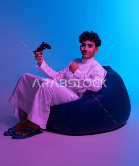 قضاء أوقات الفراغ في اللعب بالألعاب الالكترونية الترفيهية، المتعة والتسلية بالفيديو جيمز، بورتريه لشاب عربي خليجي سعودي مبتسم يرتدي الثوب التقليدي يجلس على كرسي مريح و يمسك بيده عصا التحكم ويرفع قبضته للأعلى بإيماءات الفوز، النظر إلى الكاميرا، خلفية زرقاء