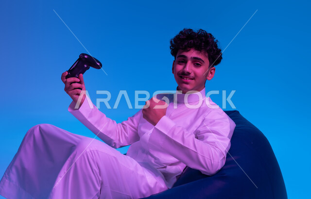 النظر إلى الكاميرا، قضاء أوقات الفراغ في اللعب بالألعاب الالكترونية الترفيهية، المتعة والتسلية بالفيديو جيمز، بورتريه لشاب عربي خليجي سعودي مبتسم يرتدي الثوب التقليدي يجلس على كرسي مريح و يمسك بيده عصا التحكم ويرفع قبضته للأعلى بإيماءات الفوز، خلفية زرقاء