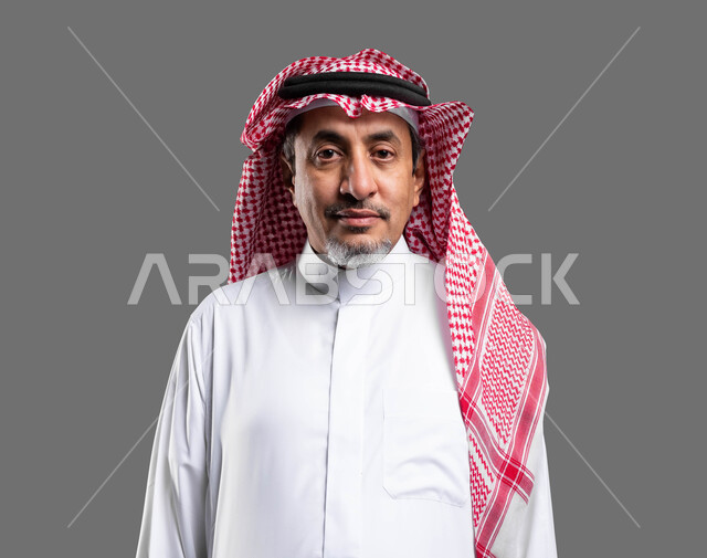 تعابير الجدية والثقة بالنفس، مفهوم الاهتمام بالمظهر الخارجي و الرجولة والأناقه، بورتريه مقرب لرجل مسن عربي خليجي سعودي مبتسم يرتدي الشماغ والثوب التقليدي يقف باستقامة وينظر إلى الكاميرا يإيماءات البشاشة، خلفية رمادية