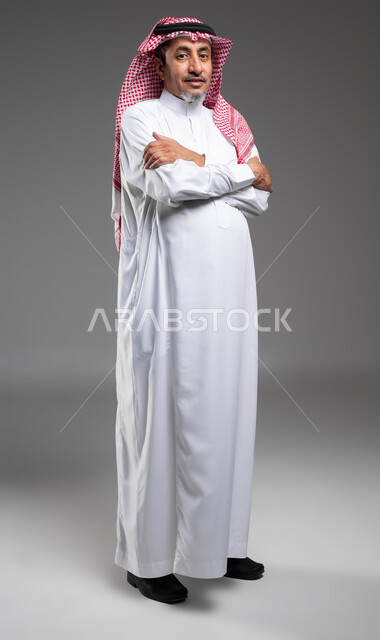 الاهتمام بالمظهر الخارجي، الوقوف بحركات أيدي متقاطعة، مفهوم الأناقة والرجولة، بورتريه لرجل كبير السن عربي خليجي سعودي مبتسم يرتدي الثوب والشماغ التقليدي ينظر إلى الكاميرا بإيماءات البرد، صورة للجسم بالكامل، خلفية رمادية.