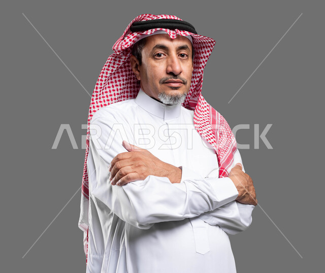 الوقوف بحركات أيدي متقاطعة، مفهوم الأناقة والرجولة، الاهتمام بالمظهر الخارجي، بورتريه مقرب لرجل كبير السن عربي خليجي سعودي مبتسم يرتدي الثوب والشماغ التقليدي ينظر إلى الكاميرا بإيماءات البرد، خلفية رمادية