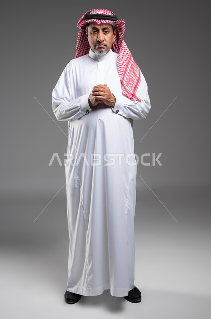 Gestures of concentration, absent-mindedness, contemplation, contemplation and planning for the future, standing with coherent hand movements, portrait of an elderly Saudi Gulf Arab man wearing traditional clothing and a shemagh, looking at the camera and thinking to find a solution to a problem, full body portrait, gray background