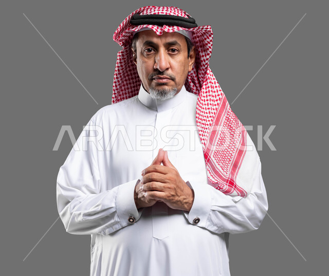 إيماءات التمعن والتركيز و شرود الذهن ،التأمل والتخطيط للمستقبل, الوقوف بحركات أيدي متماسكة، بورتريه مقرب لرجل مسن عربي خليجي سعودي يرتدي الزي التقليدي والشماغ ينظر إلى الكاميرا و يقوم بالتفكير لإيجاد حل لمشكلة ما، خلفية رمادية