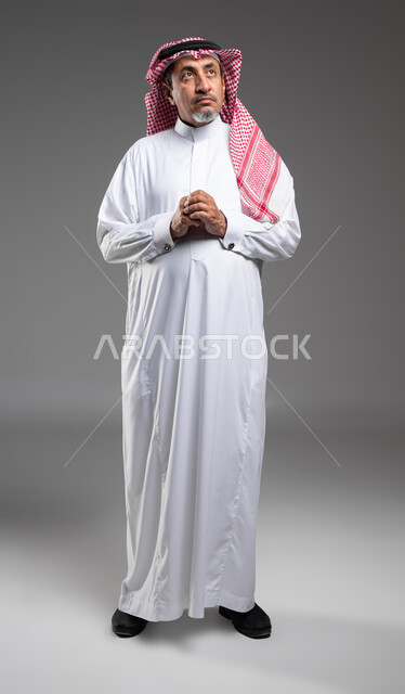 التأمل وشرود الذهن والتخطيط للمستقبل, الوقوف بحركات أيدي متماسكة، إيماءات التمعن والتركيز، بورتريه لرجل مسن عربي خليجي سعودي يرتدي الزي التقليدي والشماغ ينظر للاعلى و يقوم بالتفكير لإيجاد حل لمشكلة ما, صورة الجسم بالطول الكامل، خلفية رمادية