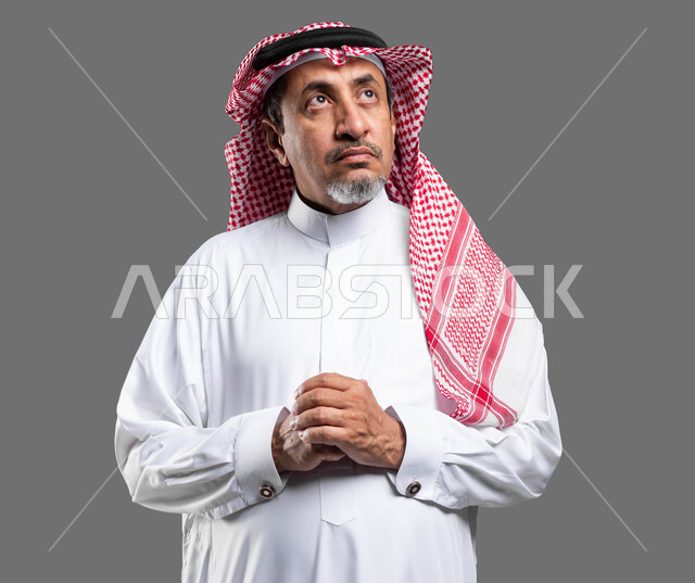 الوقوف بحركات أيدي متماسكة، إيماءات التمعن والتركيز، التأمل وشرود الذهن والتخطيط للمستقبل, بورتريه مقرب لرجل كبير السن عربي خليجي سعودي يرتدي الزي التقليدي والشماغ ينظر للاعلى و يقوم بالتفكير لإيجاد حل لمشكلة ما, خلفية رمادية