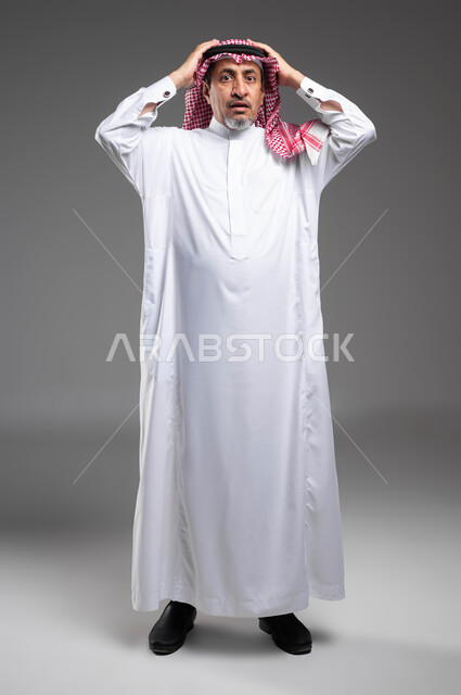 الشعور بالاستغراب والدهشة , التفاجؤ من شيء ما، الانزعاج عند سماع خبر سيء، بورتريه مقرب لرجل مسن عربي خليجي سعودي يرتدي الزي التقليدي والشماغ و يرفع يديه على رأسه بتعابير الصدمة, النظر إلى الكاميرا بإيماءات الذهول والتعجب , صورة للجسم بالكامل، خلفية رمادية