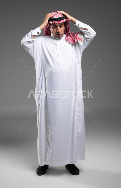 النظر إلى الاسفل بإيماءات الخسارة و الذهول ,الانزعاج عند سماع خبر سيء، التفاجؤ من شيء ما، الشعور بالدهشة والاستغراب , بورتريه لرجل مسن عربي خليجي سعودي يرتدي الزي التقليدي والشماغ و يرفع يديه على رأسه بتعابير الصدمة, صورة الجسم بالكامل، خلفية رمادية