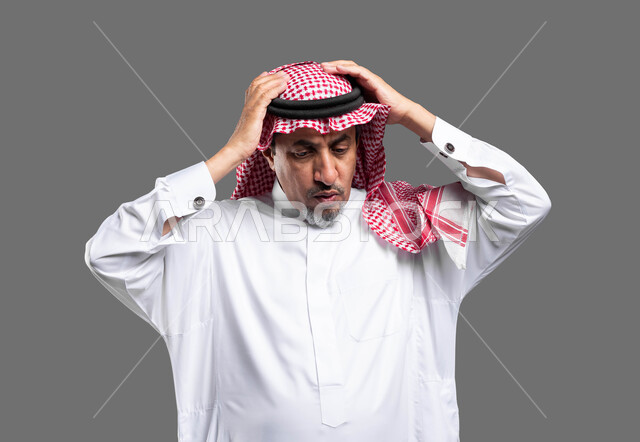 الانزعاج عند سماع خبر سيء, التفاجؤ من شيء ما، الشعور بالدهشة والاستغراب , بورتريه لرجل مسن عربي خليجي سعودي يرتدي الزي التقليدي والشماغ و يرفع يديه على رأسه بتعابير الصدمة, النظر إلى الاسفل بإيماءات الخسارة و الذهول , خلفية رمادية