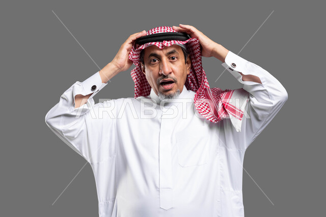 التفاجؤ من شيء ما، الانزعاج عند سماع خبر سيء، الشعور بالدهشة والاستغراب , بورتريه مقرب لرجل مسن عربي خليجي سعودي يرتدي الزي التقليدي والشماغ و يرفع يديه على رأسه بتعابير الصدمة, النظر إلى الكاميرا بإيماءات الذهول والتعجب , خلفية رمادية