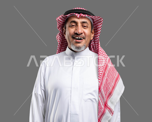 التعبير عن الفرح بالضحكات العالية، الضحك مع الفكاهات والنكت، بورتريه مقرب لرجل مسن عربي خليجي سعودي يرتدي الزي التقليدي والشماغ ينظر إلى الكاميرا بإيماءات السرور، خلفية رماديه