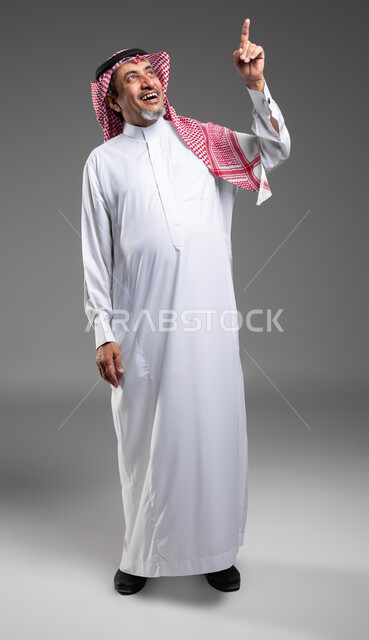 الضحك والفرح بوجود شيء مفقود، تحديد موقع معين بإيماءات تدل على السرور، إشارات وحركات تدل على هذا المكان (هنا)، بورتريه لرجل كبير السن مبتسم عربي خليجي سعودي يرتدي الزي التقليدي والشماغ يشير بأصبعه نحو الأعلى، صورة الجسم بالطول الكامل، خلفية رمادية