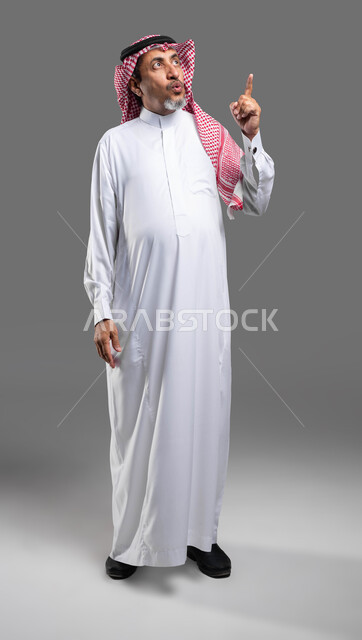 تحديد موقع معين بإيماءات فم ووجه تدل على الذهول والتعجب، إشارات وحركات تدل على هذا المكان (هنا)، بورتريه لرجل كبير السن عربي خليجي سعودي يرتدي الزي التقليدي والشماغ يشير بأصبعه نحو الأعلى، صورة الجسم بالطول الكامل، خلفية رمادية