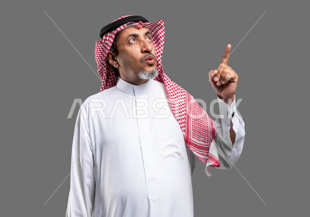 إشارات وحركات تدل على هذا المكان (هنا)، تحديد موقع معين بإيماءات فم ووجه تدل على الذهول والتعجب، بورتريه مقرب لرجل كبير السن عربي خليجي سعودي يرتدي الزي التقليدي والشماغ يشير بأصبعه نحو الأعلى، خلفية رمادية