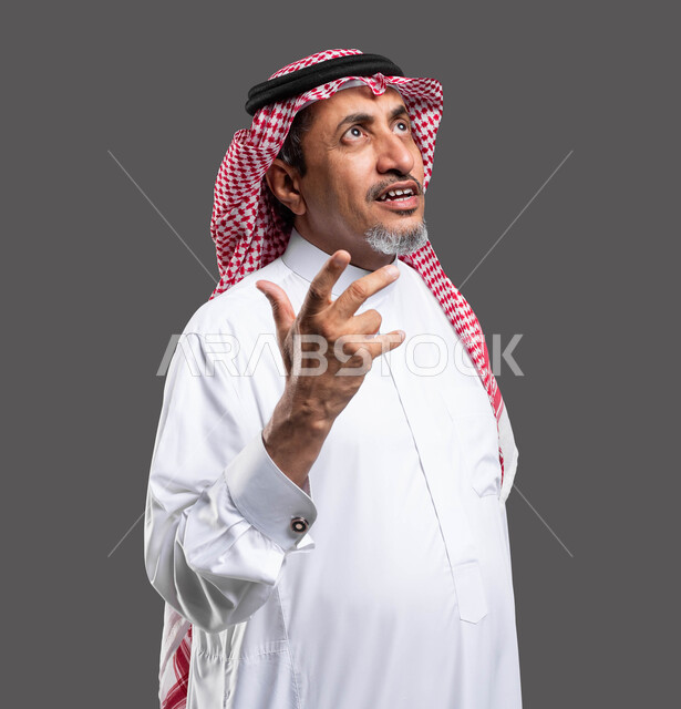 محاولة معرفة شيء ما، الاستفسار حول موضوع معين، النظر إلى الأعلى بإيماءات التكلم مع شخص ما، بورتريه مقرب لرجل كبير السن عربي خليجي سعودي يرتدي الشماغ والثوب التقليدي ويرفع يده بحركات الاستفهام، خلفية رمادية .
