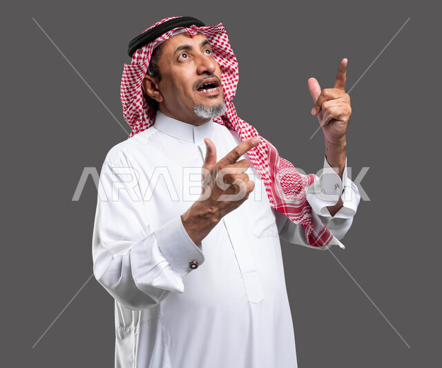 Inquiring about a specific topic, looking up with gestures of talking to someone, close-up portrait of an elderly Saudi Gulf Arab man wearing a shemagh and traditional dress and raising his hand in questioning movements, trying to find out something, gray background