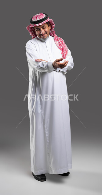 العطف والرفق والحنان، ايماءات الملاطفه والملاعبه بحنيه، بورتريه لرجل كبير السن عربي خليجي سعودي مبتسم يرتدي الشماغ والثوب التقليدي يقوم بحمل عنصر ما ، الشعور بالمحبه والاشفاق، صورة الجسم بالطول الكامل، خلفية رمادية .