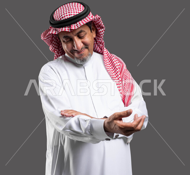 ايماءات الملاطفه والملاعبه بحنيه، العطف والرفق والحنان، بورتريه مقرب لرجل كبير السن عربي خليجي سعودي مبتسم يرتدي الشماغ والثوب التقليدي يقوم بحمل عنصر ما ،  الشعور بالمحبه والاشفاق، خلفية رمادية .