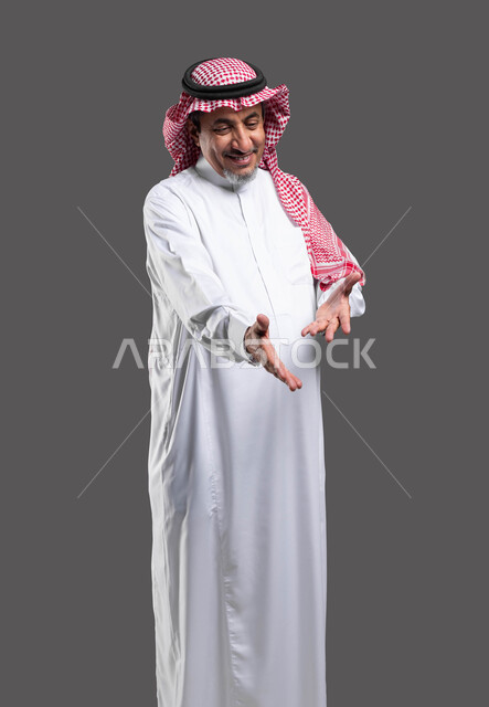 نظرات مليئة بالحب والعطف، تقديم المساعده ببشاشه وسرور، ايماءات السعاده والفرح، بورتريه مقرب لرجل كبير السن عربي خليجي سعودي مبتسم يرتدي الشماغ والثوب التقليدي يقوم بملاعبة أحد ما, خلفية رمادية .