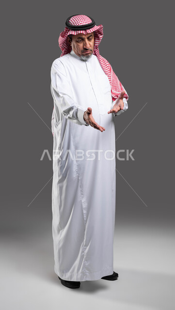 تعابير القلق وعدم الرضا، ايماءات رؤية امر محزن، عبوس الوجه والشعور بالاستياء، بورتريه لرجل كبير السن عربي خليجي سعودي مبتسم يرتدي الشماغ والثوب التقليدي يقوم بالنظر والإشارة إلى شيء ما, صورة الجسم بالطول الكامل، خلفية رمادية .