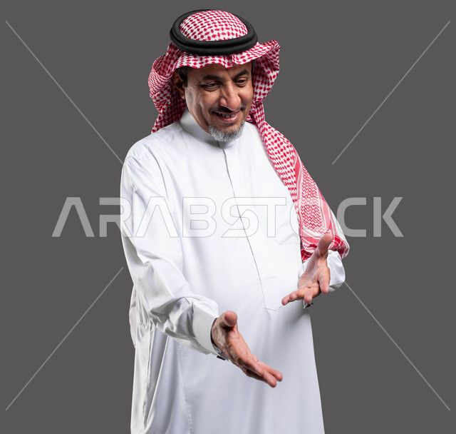 تقديم المساعده ببشاشه وسرور، ايماءات السعاده والفرح، بورتريه مقرب لرجل كبير السن عربي خليجي سعودي مبتسم يرتدي الشماغ والثوب التقليدي يقوم بملاعبة أحد ما, نظرات مليئة بالحب والعطف، خلفية رمادية .