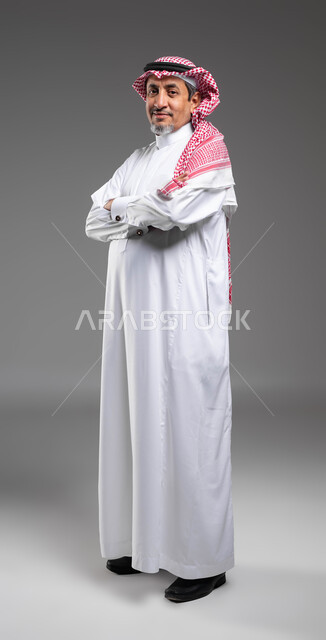الاهتمام بالأناقة والمظهر الخارجي, مفهوم الرجولة والثقة بالنفس، بورتريه لرجل كبير السن عربي خليجي سعودي مبتسم يرتدي الشماغ والثوب التقليدي يقف بحركات ايدي متقاطعة ينظر إلى الكاميرا بإيماءات السرور, صورة الجسم بالطول الكامل، خلفية رمادية .