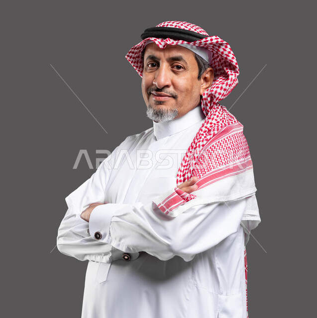 مفهوم الرجولة والثقة بالنفس، الاهتمام بالأناقة والمظهر الخارجي, بورتريه مقرب لرجل كبير السن عربي خليجي سعودي مبتسم يرتدي الشماغ والثوب التقليدي يقف بحركات ايدي متقاطعة ينظر إلى الكاميرا بإيماءات السرور,  خلفية رمادية .