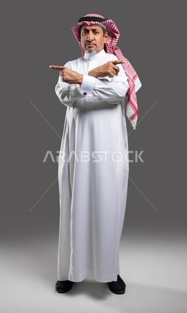 تعابير الجدية والحزم، الإشارة إلى طرفين متعاكسين, بورتريه كبير السن لرجل عربي خليجي سعودي يرتدي الشماغ والثوب التقليدي ينظر إلى الكاميرا بإيماءات الغضب, الوقوف باستقامة بحركات أيدي متقاطعة, الشعور بالتهيج والتوتر، صورة الجسم بالطول الكامل، خلفية رمادية .