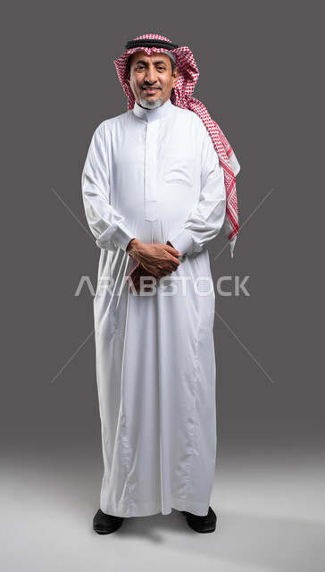 مفهوم الثقة بالنفس، الاهتمام بالأناقة والمظهر الخارجي, بورتريه لرجل كبير السن عربي خليجي سعودي مبتسم يرتدي الشماغ والثوب التقليدي يقف باستقامة بحركات ايدي متماسكة ينظر إلى الكاميرا بإيماءات السرور, صورة الجسم بالطول الكامل، خلفية رمادية .