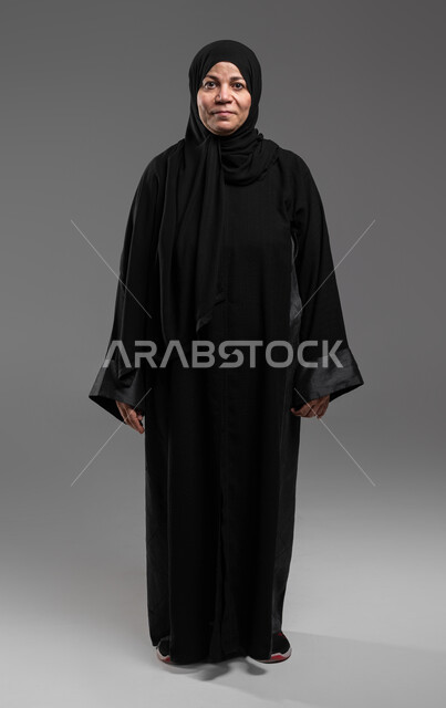 الاهتمام بالأناقة والمظهر الخارجي، إيماءات الفرح والسعادة، بورتريه لإمرأة كبيرة السن عربية خليجية سعودية مبتسمة ترتدي العباءة السوداء تنظر إلى الكاميرا بتعابير الاطمئنان ، التأمل والنظر إلى الأشياء السارة، خلفية رمادية .