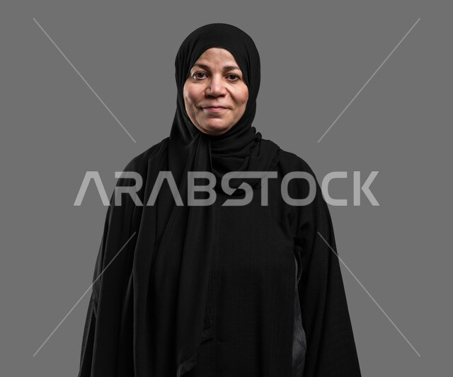 إيماءات الفرح والسعادة، الاهتمام بالأناقة والمظهر الخارجي، بورتريه مقرب لإمرأة عربية خليجية سعودية مبتسمة ترتدي العباءة السوداء تنظر إلى الكاميرا بتعابير الاطمئنان ، التأمل والنظر إلى الأشياء السارة، خلفية رمادية .