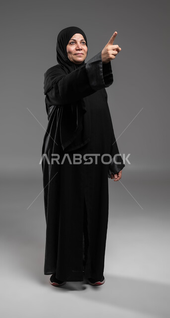 إيماءات السرور والاطمئنان برؤية الأشياء السارة، التأمل والنظر إلى حدث ما، بورتريه لإمرأة عربية خليجية سعودية مبتسمة ترتدي العباءة السوداء ترفع يدها نحو الأعلى للإشارة إلى شيء ما، صورة الجسم بالطول الكامل، خلفية رمادية .