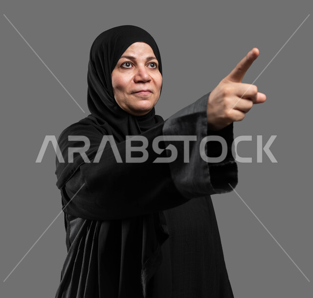 التأمل والنظر إلى حدث ما، بورتريه مقرب لإمرأة عربية خليجية سعودية مبتسمة ترتدي العباءة السوداء ترفع يدها نحو الأعلى للإشارة إلى شيء ما، إيماءات السرور والاطمئنان برؤية الأشياء السارة، خلفية رمادية .