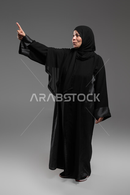 إيماءات الذهول والدهشة، استخدام حاسة اللمس لتحريك العناصر الهولوجرامية، الشعور بالمفاجأة والصدمة، بورتريه لإمرأة عربية خليجية سعودية ترتدي العباءة السوداء ترفع يدها نحو الأعلى للإشارة إلى شيء ما، صورة الجسم بالطول الكامل، خلفية رمادية .