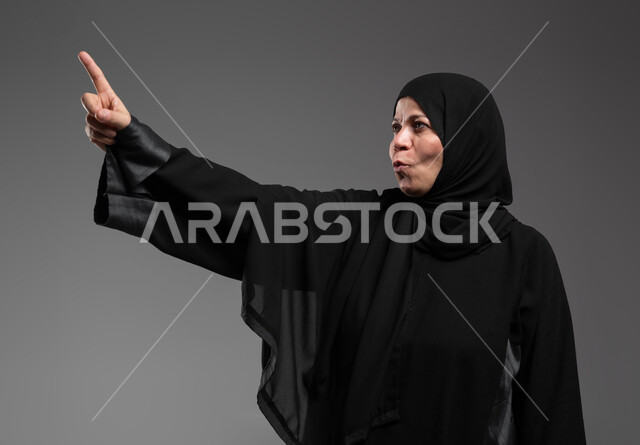 استخدام حاسة اللمس لتحريك العناصر الهولوجرامية، إيماءات الدهشة والذهول، الشعور بالمفاجأة والصدمة، بورتريه مقرب لإمرأة عربية خليجية سعودية ترتدي العباءة السوداء ترفع يدها نحو الأعلى للإشارة إلى شيء ما، خلفية رمادية .