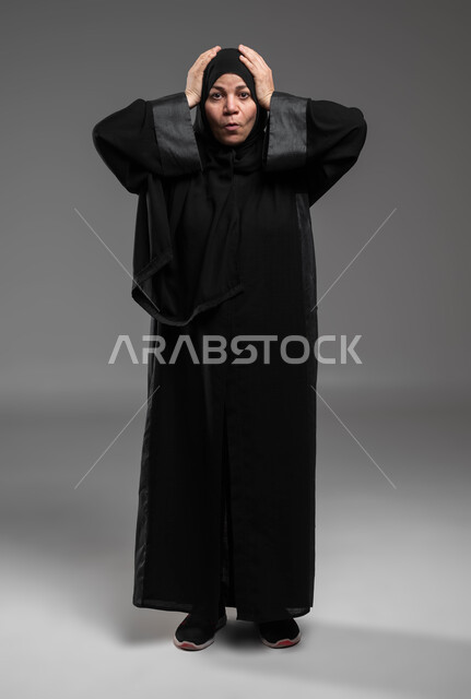 النظر إلى الكاميرا بإيماءات الدهشة والذهول، الشعور بالمفاجأة والصدمة، الانزعاج والاستغراب من شيء ما، بورتريه لإمرأة عربية خليجية سعودية ترتدي العباءة السوداء تضع يديها على رأسها تعجبا بحدوث أمر ما، صورة الجسم بالطول الكامل، خلفية رمادية .
