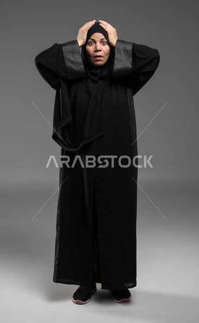 الانزعاج بسماع الأخبار السيئة، النظر إلى الكاميرا بإيماءات الدهشة والذهول، الشعور بالمفاجأة والصدمة، بورتريه لإمرأة عربية خليجية سعودية ترتدي العباءة السوداء تضع يديها على رأسها تعجبا بحدوث أمر ما، صورة الجسم بالطول الكامل، خلفية رمادية .