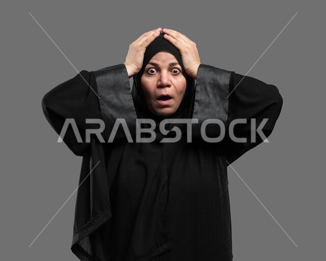 إيماءات الدهشة والذهول، الانزعاج بسماع الأخبار السيئة، الشعور بالمفاجأة والصدمة، بورتريه مقرب لإمرأة عربية خليجية سعودية ترتدي العباءة السوداء تنظر إلى الكاميرا تضع يديها على رأسها تعجبا بحدوث أمر ما، خلفية رمادية .
