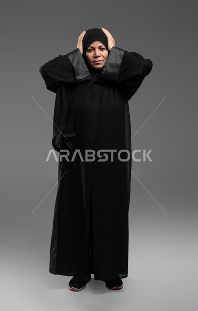الشعور بالمفاجأة والصدمة، الانزعاج والاستغراب من شيء ما، بورتريه لإمرأة عربية خليجية سعودية ترتدي العباءة السوداء تضع يديها على رأسها تعجبا بحدوث أمر ما، النظر إلى الكاميرا بإيماءات الدهشة والذهول، صورة الجسم بالطول الكامل، خلفية رمادية .