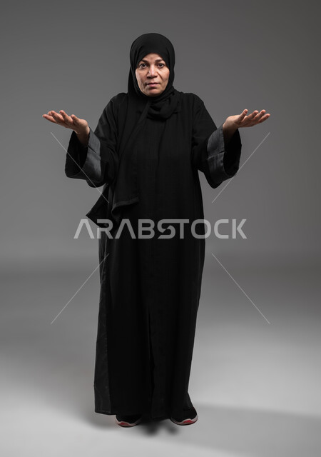 الشعور بالانزعاج والاستغراب، النظر إلى الكاميرا بإيماءات الاستفهام والاستفسار، بورتريه لإمرأة عربية خليجية سعودية ترتدي العباءة السوداء ترفع يديها بكفوف مفتوحة نحو الأعلى للسؤال عن أمر ما، موضوع مبهم و مجهول، صورة الجسم بالطول الكامل، خلفية رمادية .