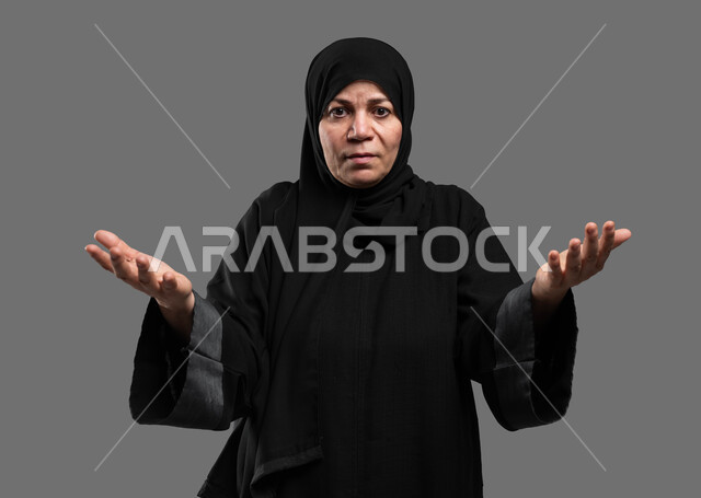 النظر إلى الكاميرا بإيماءات الاستفهام والاستفسار، بورتريه مقرب لإمرأة عربية خليجية سعودية ترتدي العباءة السوداء ترفع يديها بكفوف مفتوحة نحو الأعلى للسؤال عن أمر ما، موضوع مبهم و مجهول، الشعور بالانزعاج والاستغراب، خلفية رمادية .