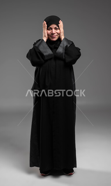 الشعور بالسعادة والسرور بسماع الأخبار السارة، النظر إلى الكاميرا بإيماءات المفاجأة والذهول, بورتريه لإمرأة عربية سعودية خليجية محجبة تضع يديها على رأسها بتعابير الدهشة, الإستغراب بحدوث أمر ما، صورة الجسم بالطول الكامل، خلفية رمادية .