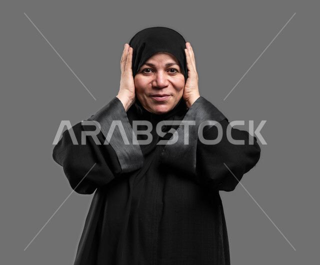 إيماءات المفاجأة والذهول, الشعور بالسعادة والسرور بسماع الأخبار السارة، بورتريه مقرب لإمرأة عربية سعودية خليجية محجبة تنظر إلى الكاميرا  تضع يديها على رأسها بتعابير الدهشة, الإستغراب بحدوث أمر ما ، خلفية رمادية .