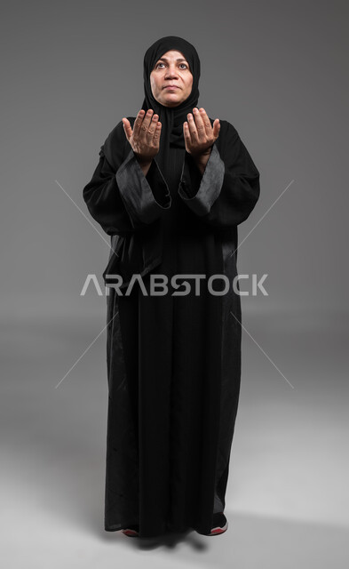 إيماءات التمني والطلب, العبادة والتقرب إلى الله، بورتريه لإمرأة عربية سعودية خليجية محجبة ترفع يديها للأعلى للدعاء, أدعية وأذكار، طلب المغفرة وتحقيق الأمنيات،الاجتهاد في الطاعات، صورة الجسم بالطول الكامل، خلفية رمادية .