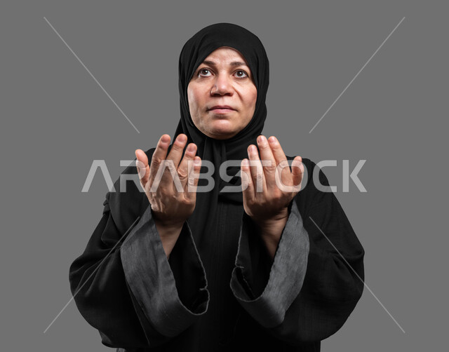 العبادة والتقرب إلى الله، إيماءات التمني والطلب, بورتريه مقرب لإمرأة عربية سعودية خليجية محجبة ترفع يديها للأعلى للدعاء, أدعية وأذكار، طلب المغفرة وتحقيق الأمنيات،الاجتهاد في الطاعات، خلفية رمادية .