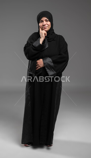 شرود الذهن في موضوع معين، النظر إلى الكاميرا بإيماءات التأمل والتمعن, تعابير الاندماج و الحيرة , بورتريه لإمرأة عربية سعودية خليجية محجبة تضع يدها تحت رأسها وتفكر, إيجاد الحلول لمشكلة ما، صورة الجسم بالطول الكامل، خلفية رمادية .