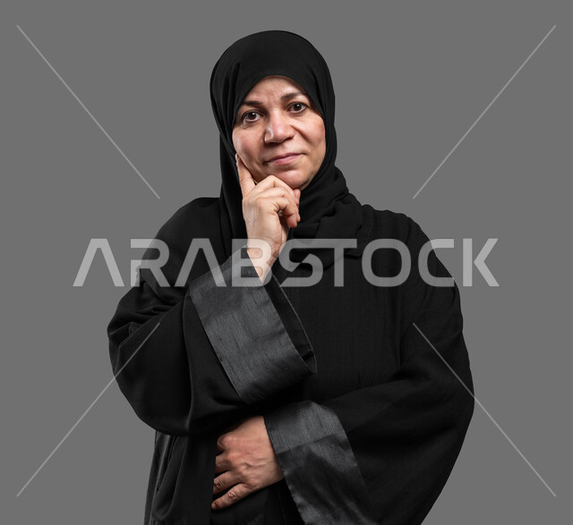 إيماءات التأمل والتمعن, النظر إلى الكاميرا بتعابير الاندماج و الحيرة , بورتريه مقرب لإمرأة عربية سعودية خليجية محجبة تضع يدها تحت رأسها وتفكر, شرود الذهن في موضوع معين، إيجاد الحلول لمشكلة ما، خلفية رمادية .