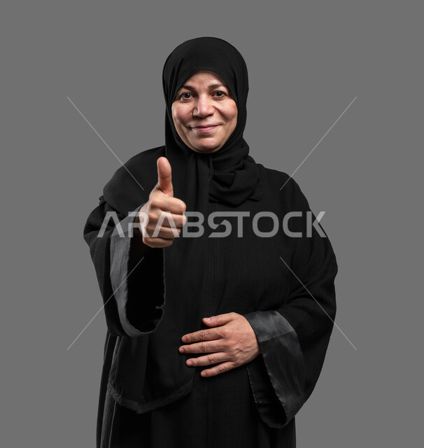 إيماءات الإعجاب والرضا، الشعور بالقبول والنظر إلى الكاميرا، بورتريه مقرب لسيدة عربية خليجية سعودية مبتسمة محجبة كبيرة بالسن مبتسمة ترتدي العباءة السوداء تقف ترفع إصبع الإبهام نحو الأعلى بعلامة الجودة، خلفية رمادية.